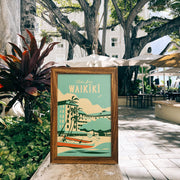Framed poster of Waikiki with a tropical scene, placed outdoors near trees and plants.
