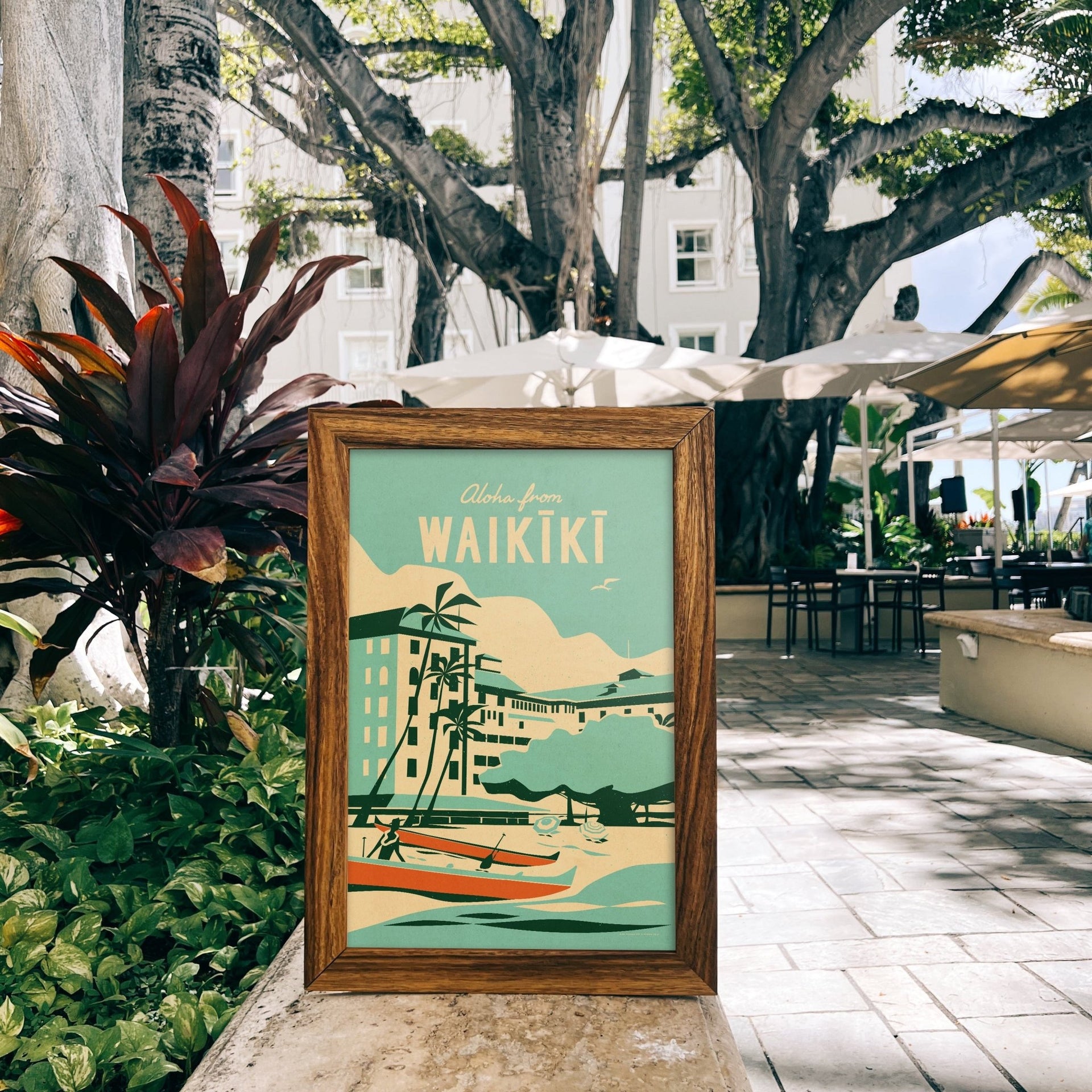 Framed poster of Waikiki with a tropical scene, placed outdoors near trees and plants.