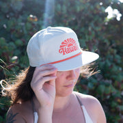 Person wearing a white cap with 'Hawaii' printed on it, standing against a green leafy background.