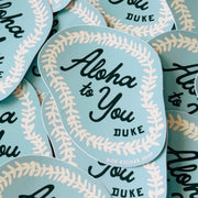 A close-up of multiple matching stickers arranged overlapping. Each sticker is light blue with a cream floral lei pattern border and black script text reading 'Aloha to You DUKE.