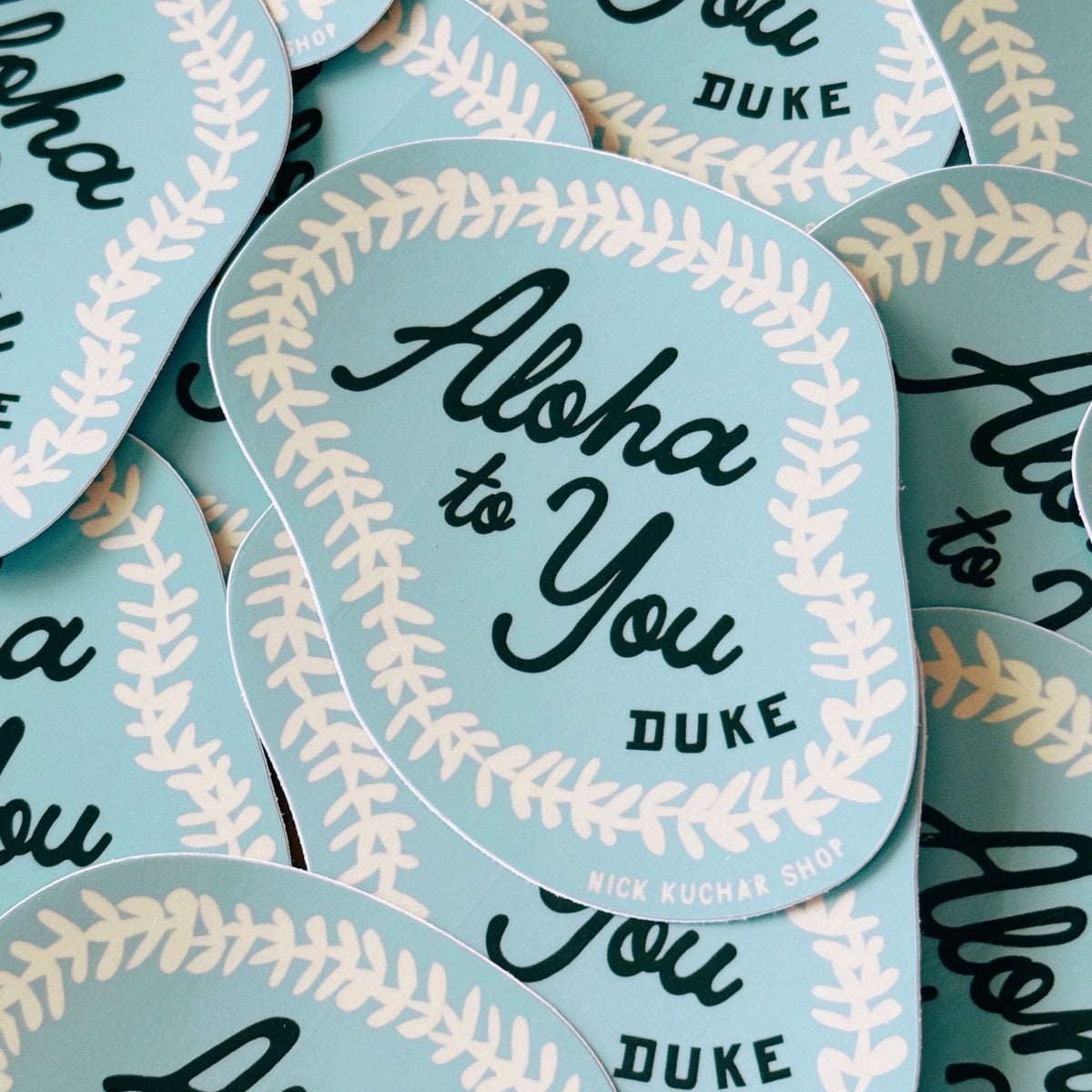 A close-up of multiple matching stickers arranged overlapping. Each sticker is light blue with a cream floral lei pattern border and black script text reading 'Aloha to You DUKE.