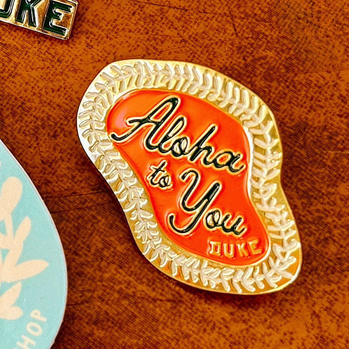 A close-up of an enamel pinwith a gold floral lei border, an orange-red center, and the words 'Aloha to You DUKE' in black script and block lettering, placed on a brown textured surface.