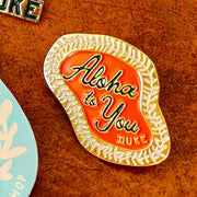 A close-up of an enamel pinwith a gold floral lei border, an orange-red center, and the words 'Aloha to You DUKE' in black script and block lettering, placed on a brown textured surface.