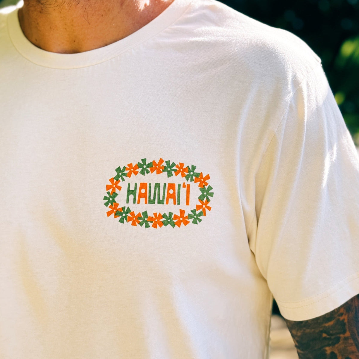 A close-up of a man wearing a cream-colored t-shirt with a graphic on the chest. The graphic features the word 'HAWAII' in green letters, framed by orange flowers and green leaves in a floral oval.