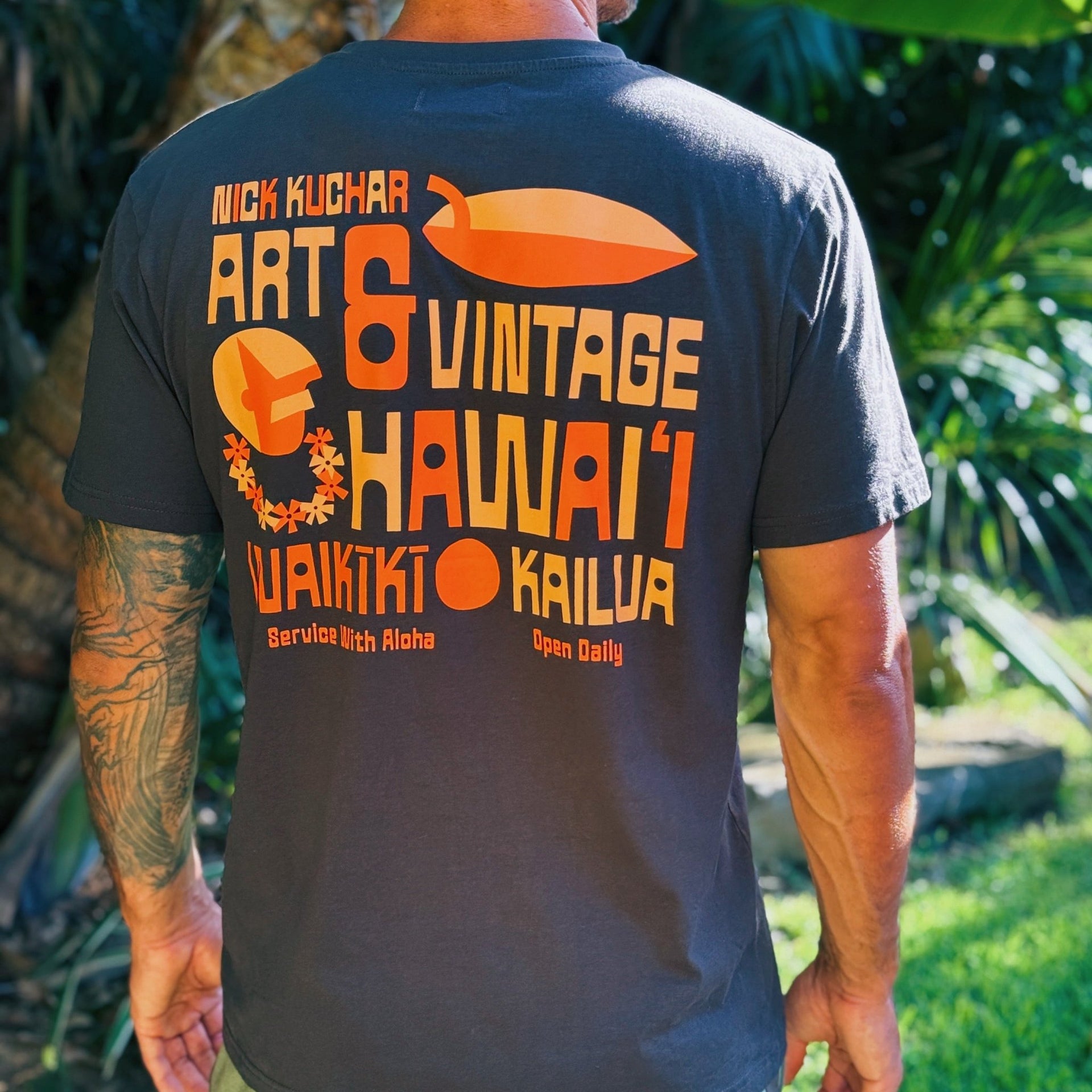 A man viewed from behind wearing a dark charcoal gray t-shirt with a large orange and yellow graphic print on the back. The print features Hawaiian text and art elements.