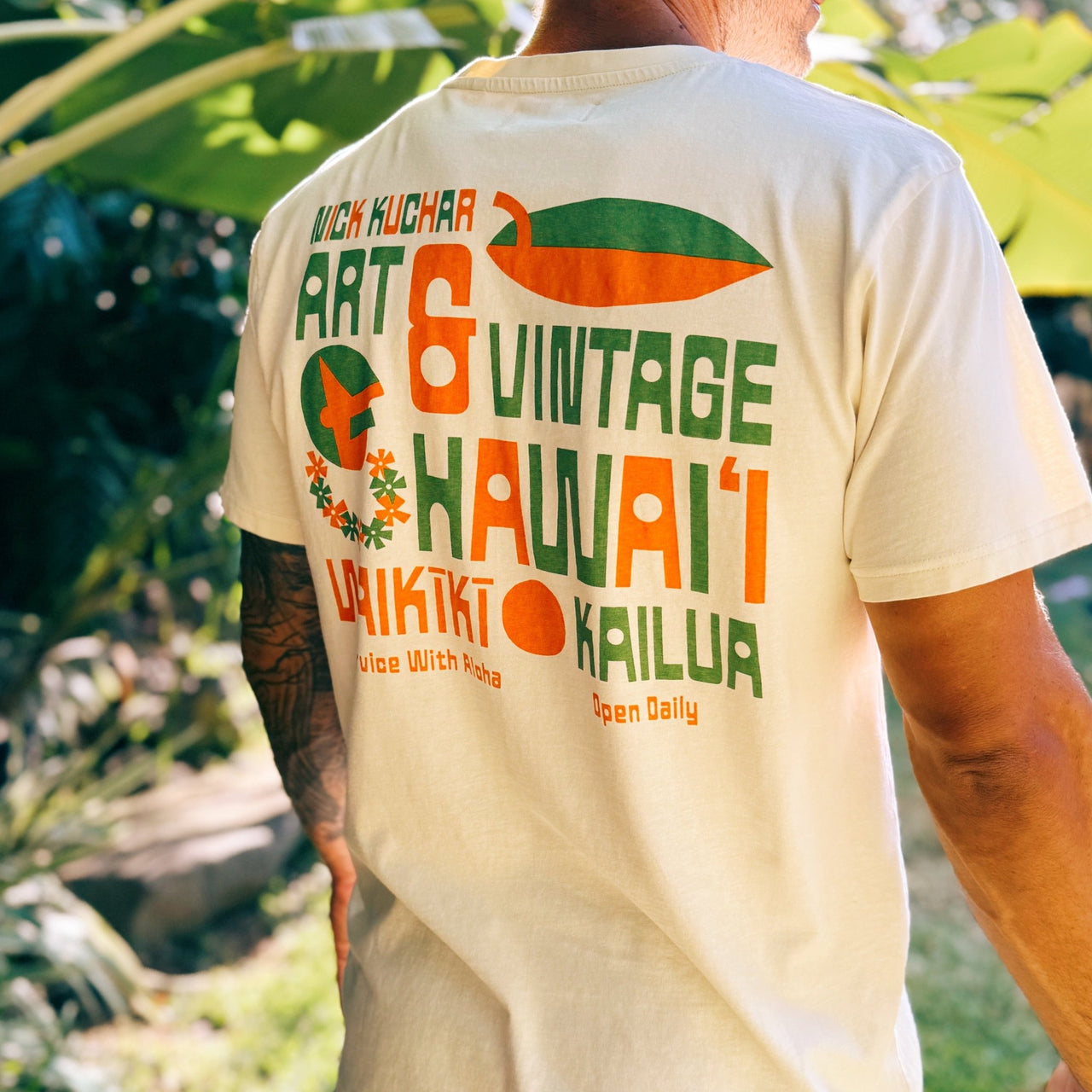A man wearing a cream-colored t-shirt with a large green and orange graphic print on the back, standing outdoors in a garden setting.