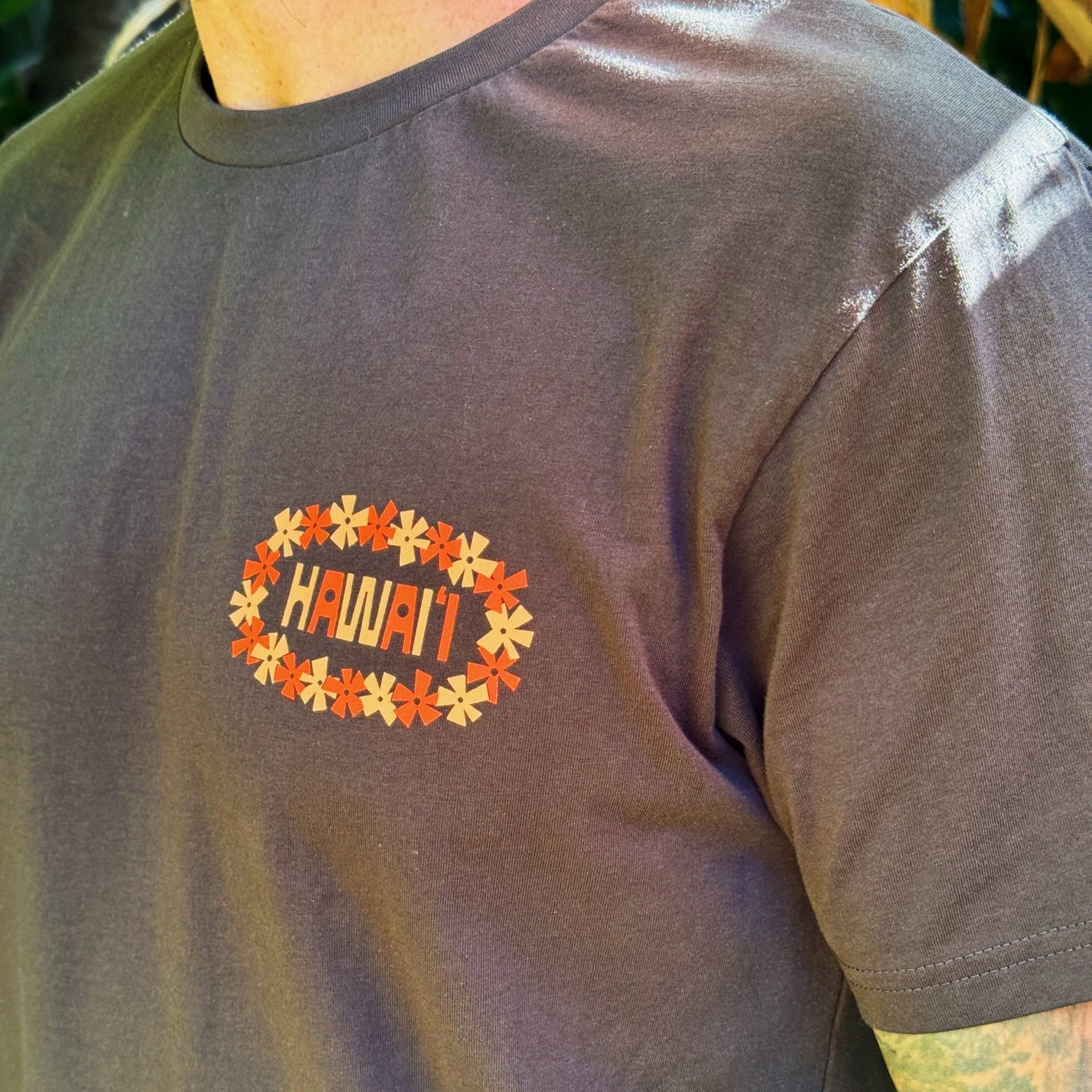 A close-up of a person wearing a dark charcoal gray t-shirt with a 'HAWAII' graphic on the chest, surrounded by a floral wreath design.