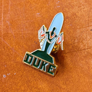 A close-up of a metal enamel pin featuring a light blue duke statue on a gold with the word 'DUKE' in deark letters outlined in gold, placed on a textured orange-brown surface.