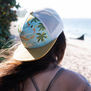 Person wearing a colorful cap with palm trees and fish design, standing by a body of water.