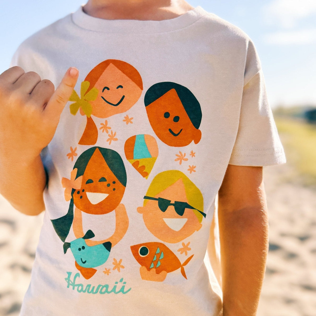 Child wearing a t-shirt with colorful graphics of children and fish, with 'Hawaii' text, on a beach.