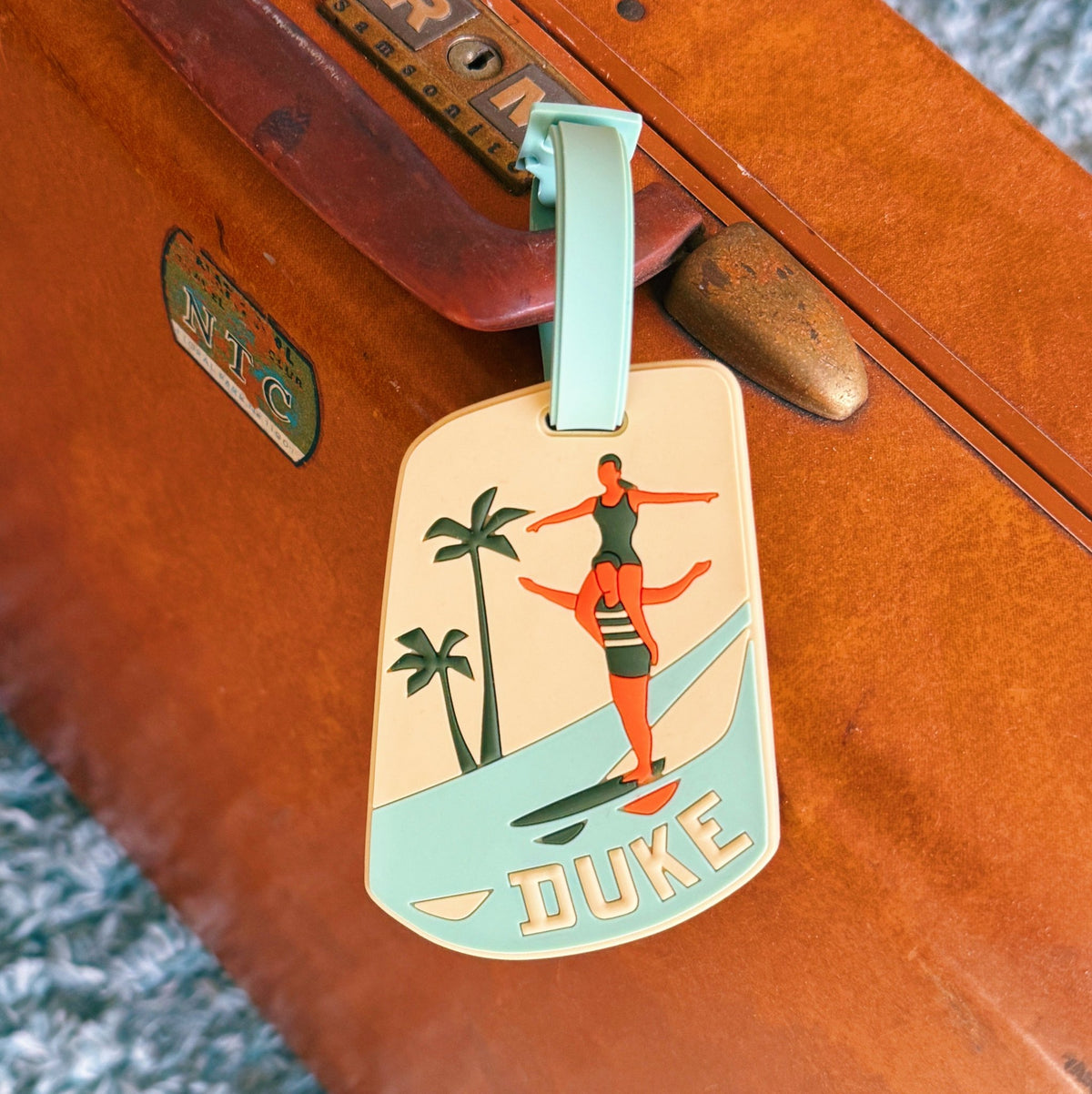 A vintage-style dog tag luggage tag with a mint green loop, featuring retro artwork of a woman balancing on a surfboard with palm trees and the word 'DUKE' at the bottom, shown attached to a brown suitcase.