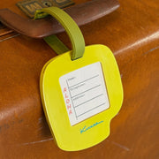 A bright yellow luggage tag with an olive green strap attached to a brown wooden suitcase. The tag features a white insert with fields for name, destination, and address, along with the word 'ALOHA' printed vertically and a blue signature logo at the bottom.
