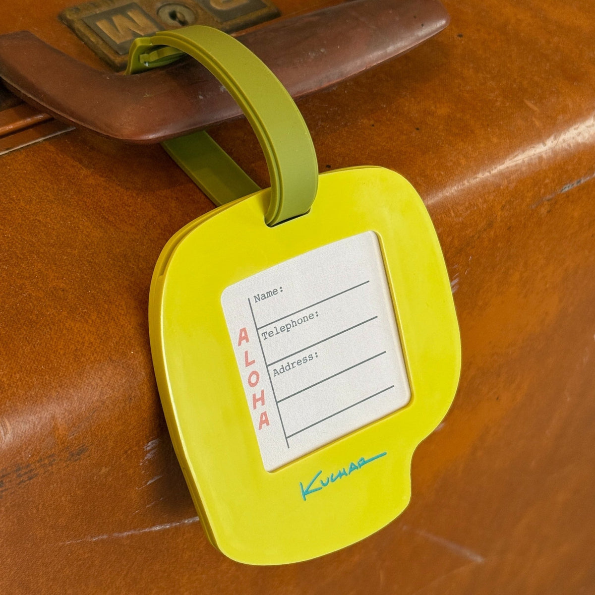 A bright yellow luggage tag with an olive green strap attached to a brown wooden suitcase. The tag features a white insert with fields for name, destination, and address, along with the word 'ALOHA' printed vertically and a blue signature logo at the bottom.