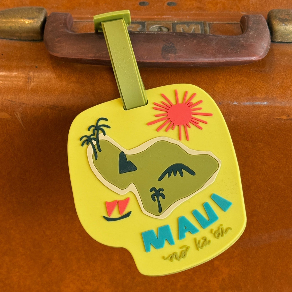 A yellow luggage tag featuring a colorful illustration of a tropical island with palm trees, a red sun, and the word 'MAUI' in blue, hanging from a green strap on a brown suitcase.