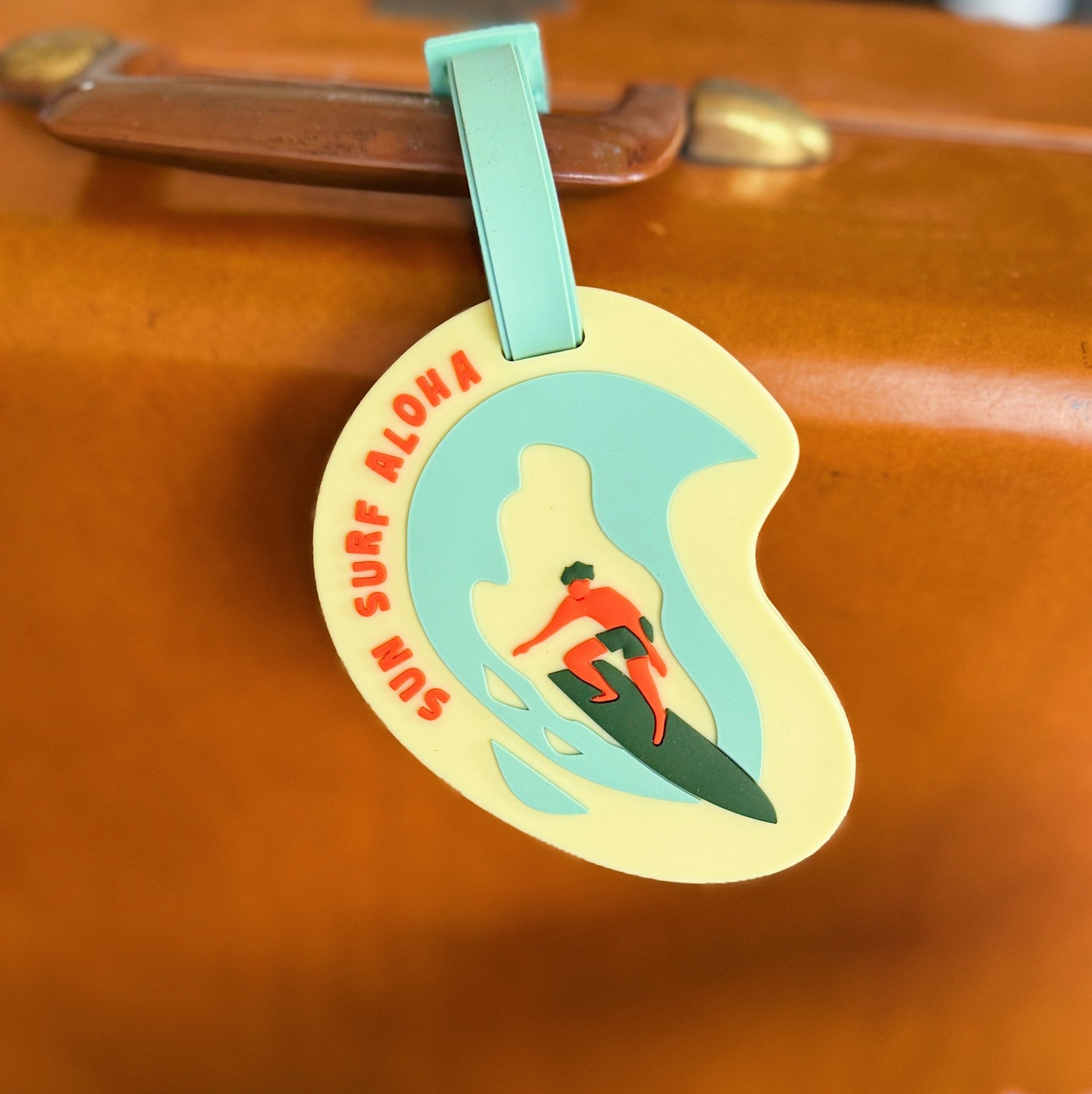 Luggage tag with a surf design and 'Sun Surf Aloha' text on a brown suitcase.