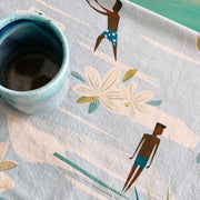 A close-up of a light blue kitchen towel with a printed illustration of two men in swim trunks, white flowers, and abstract beach-like shapes. A ceramic cup sits on the towel