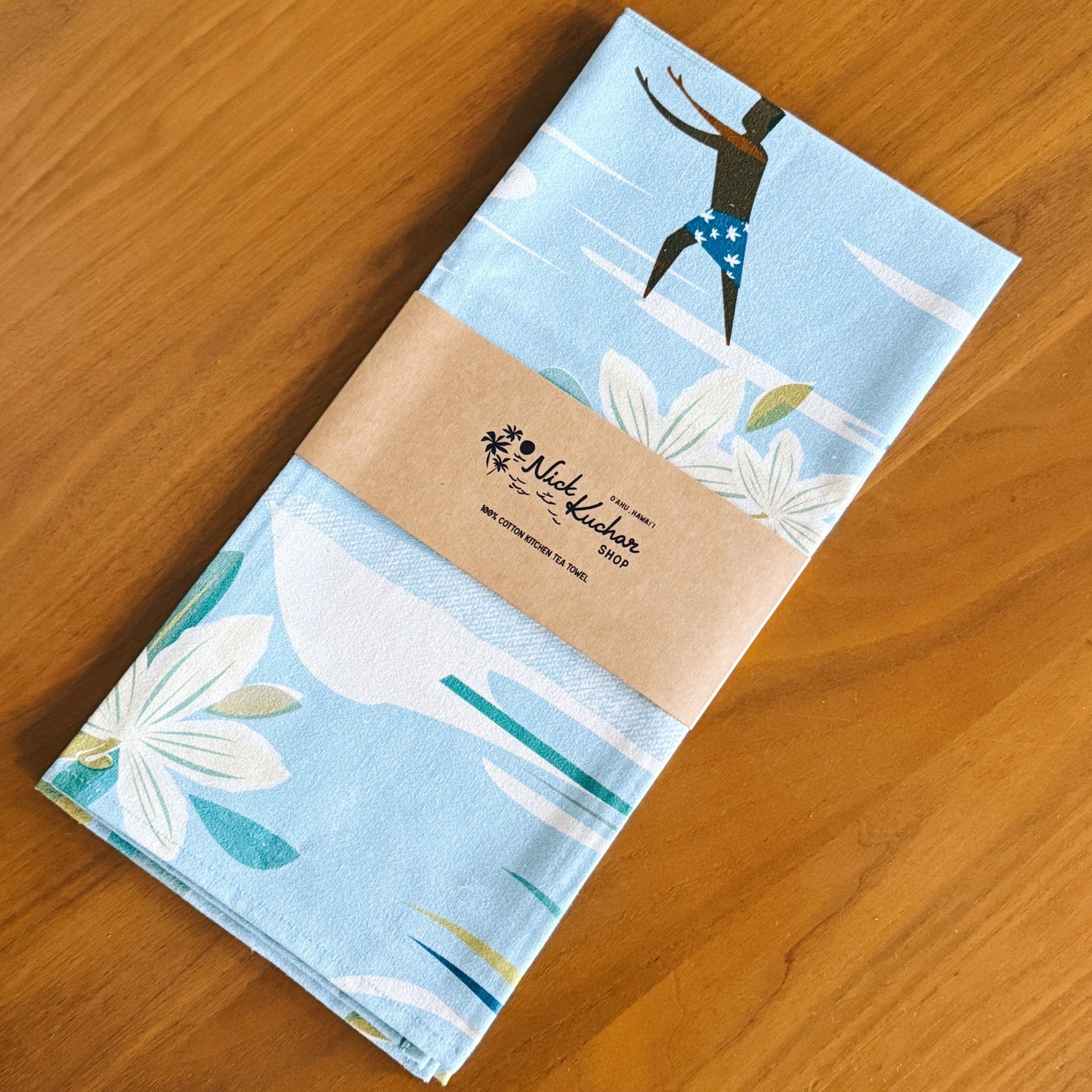 A folded light blue kitchen towel with a white floral print and a black-and-blue diver illustration, wrapped in a kraft paper band and placed on a wooden table.