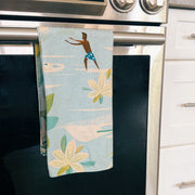 A kitchen towel with a tropical print of a surfer and palm leaves hanging from the handle of a stainless steel oven.