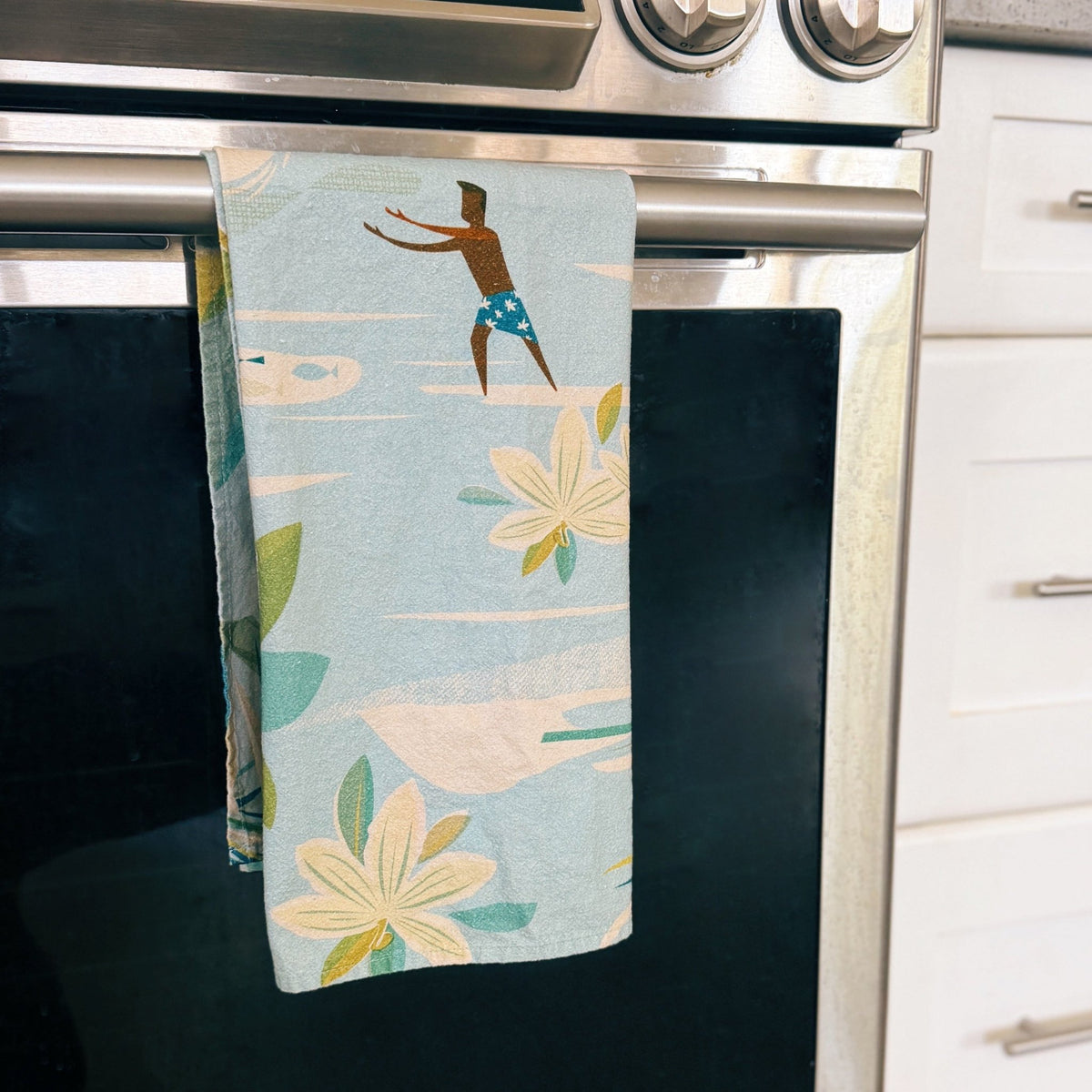 A kitchen towel with a tropical print of a surfer and palm leaves hanging from the handle of a stainless steel oven.