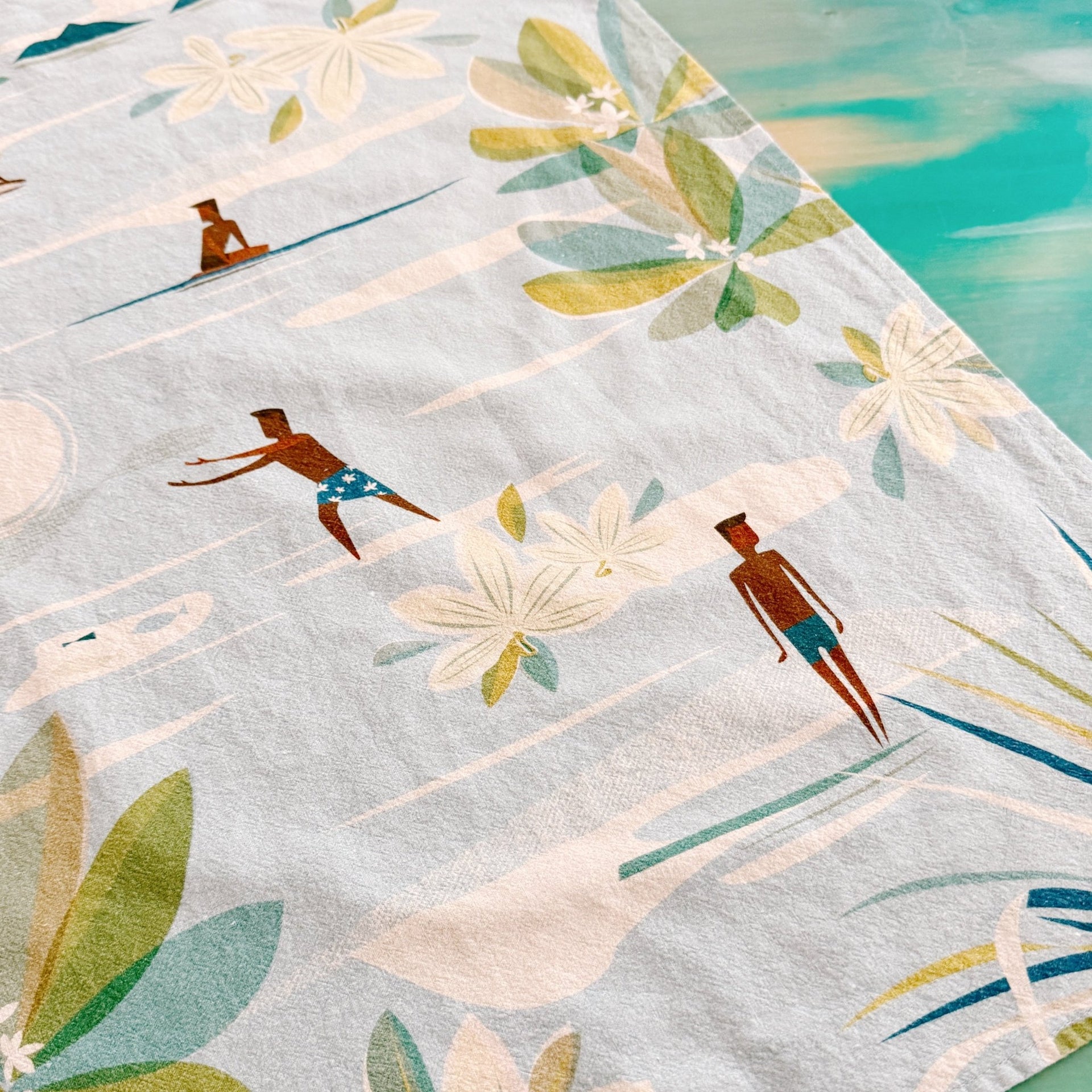 A light blue beach towel featuring a retro-style tropical print with palm leaves, white flowers, and figures of people sunbathing on the beach.