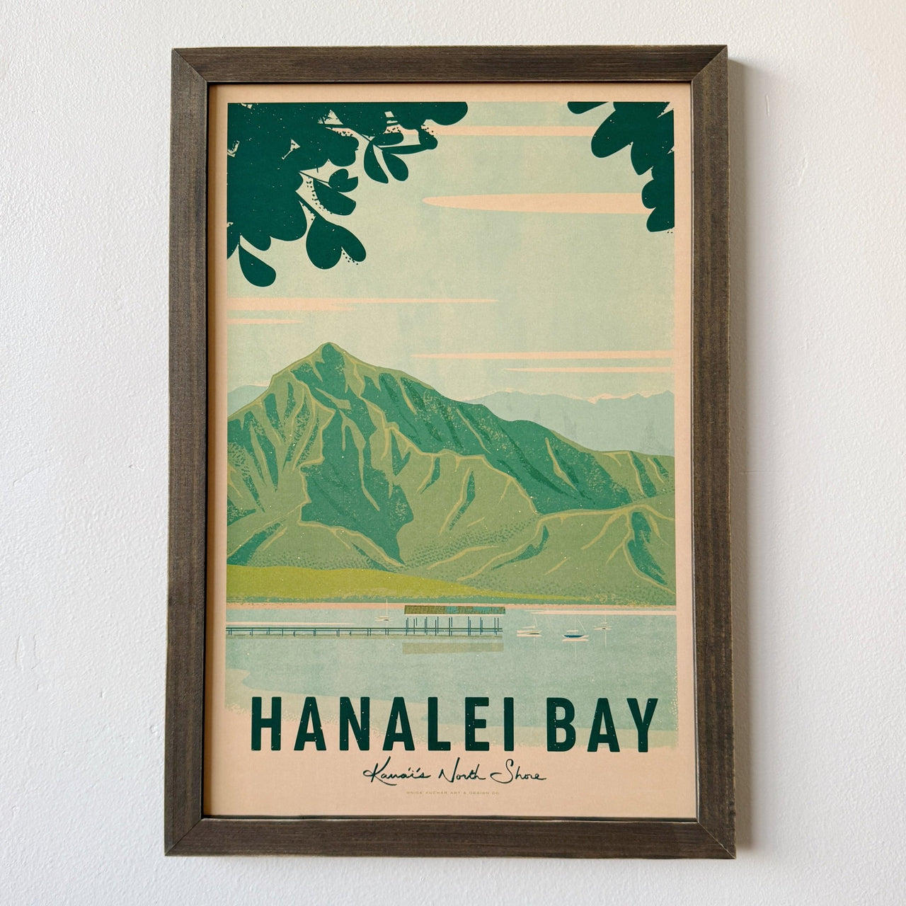 A framed travel poster of Hanalei Bay, Hawaii, featuring a retro-style illustration of green mountains, a pier, and a boat on a light blue and beige background, mounted on a white wall.