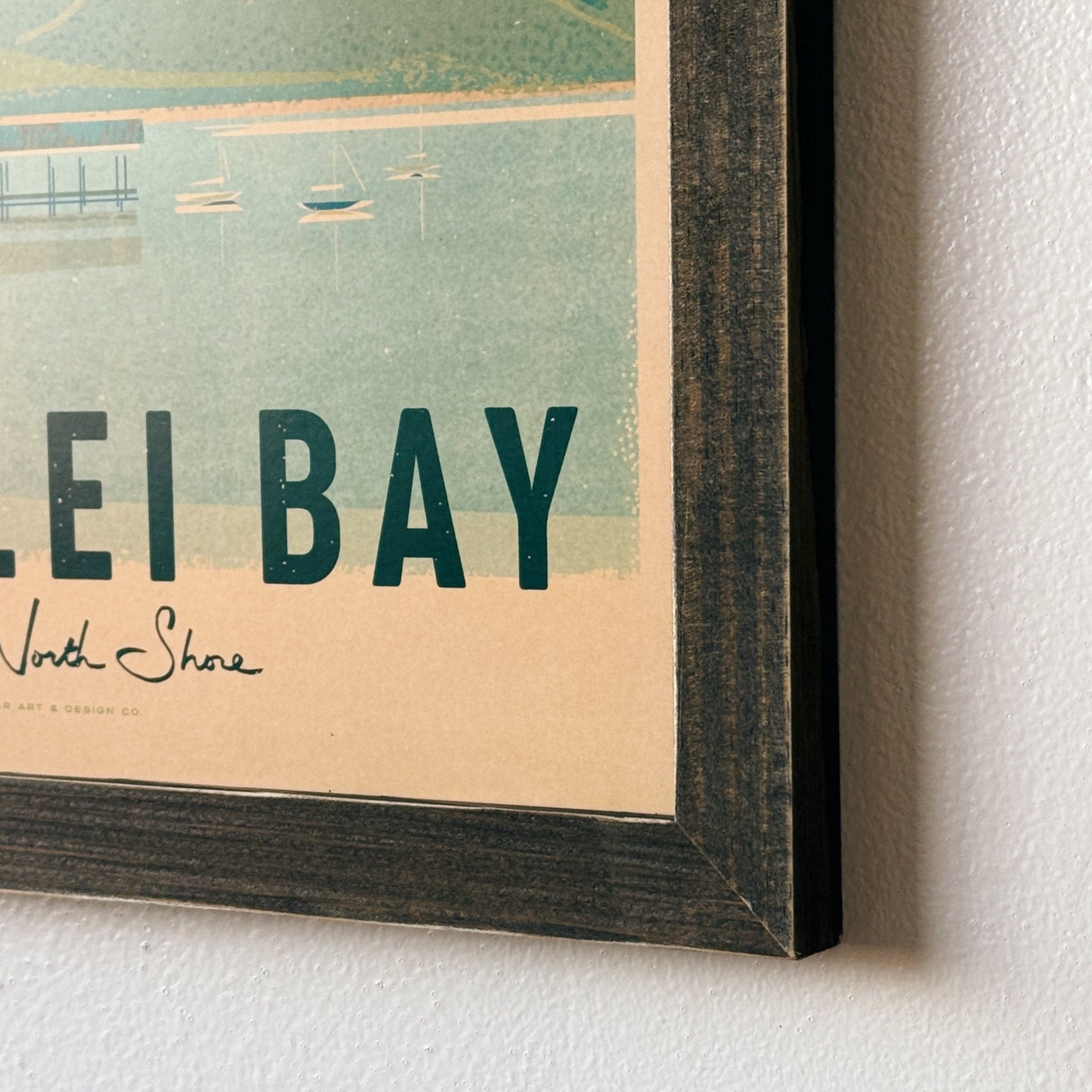 A framed vintage-style travel poster for Lei Bay, North Shore, hanging on a white textured wall. The poster features a teal and beige illustration with the text 'LEI BAY' and 'North Shore', shown in a dark wood frame.