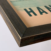 Close-up of a framed art print showing a dark wood frame corner with a vintage-style scenic print featuring green landscape elements and partial text on a beige background.
