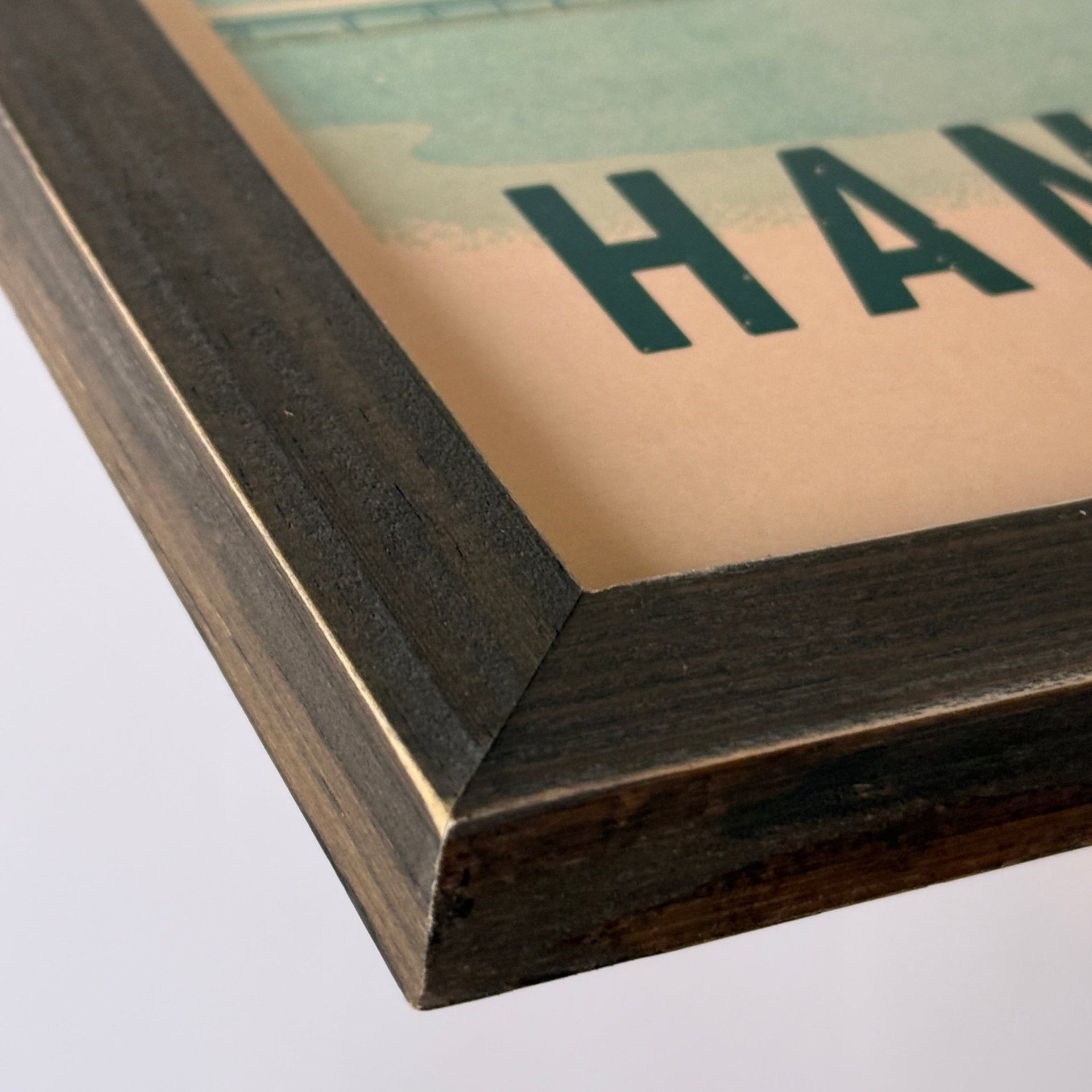 Close-up of a framed art print showing a dark wood frame corner with a vintage-style scenic print featuring green landscape elements and partial text on a beige background.
