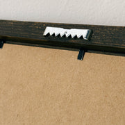 Close-up of the back side of a framed picture, showing a dark wood frame, a silver metal mounting bracket, and a brown cardboard backing against a white wall.