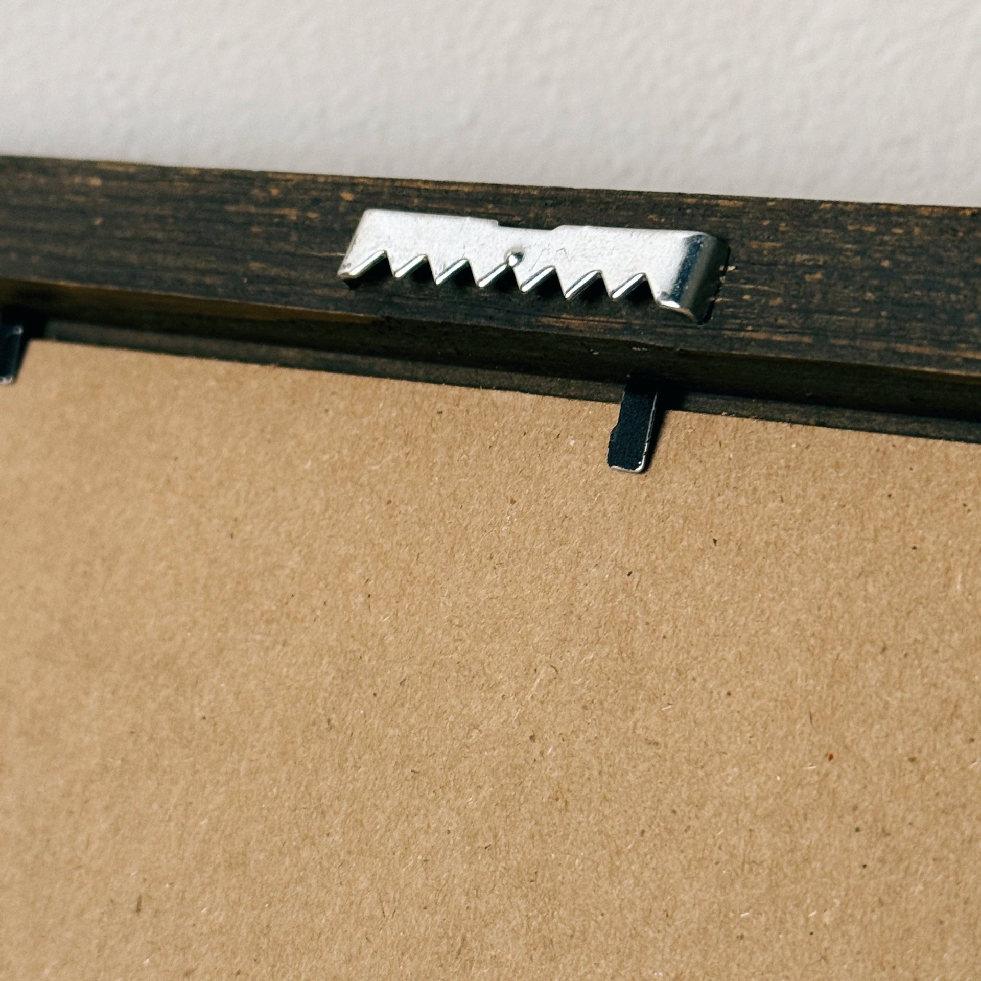 Close-up of the back side of a framed picture, showing a dark wood frame, a silver metal mounting bracket, and a brown cardboard backing against a white wall.