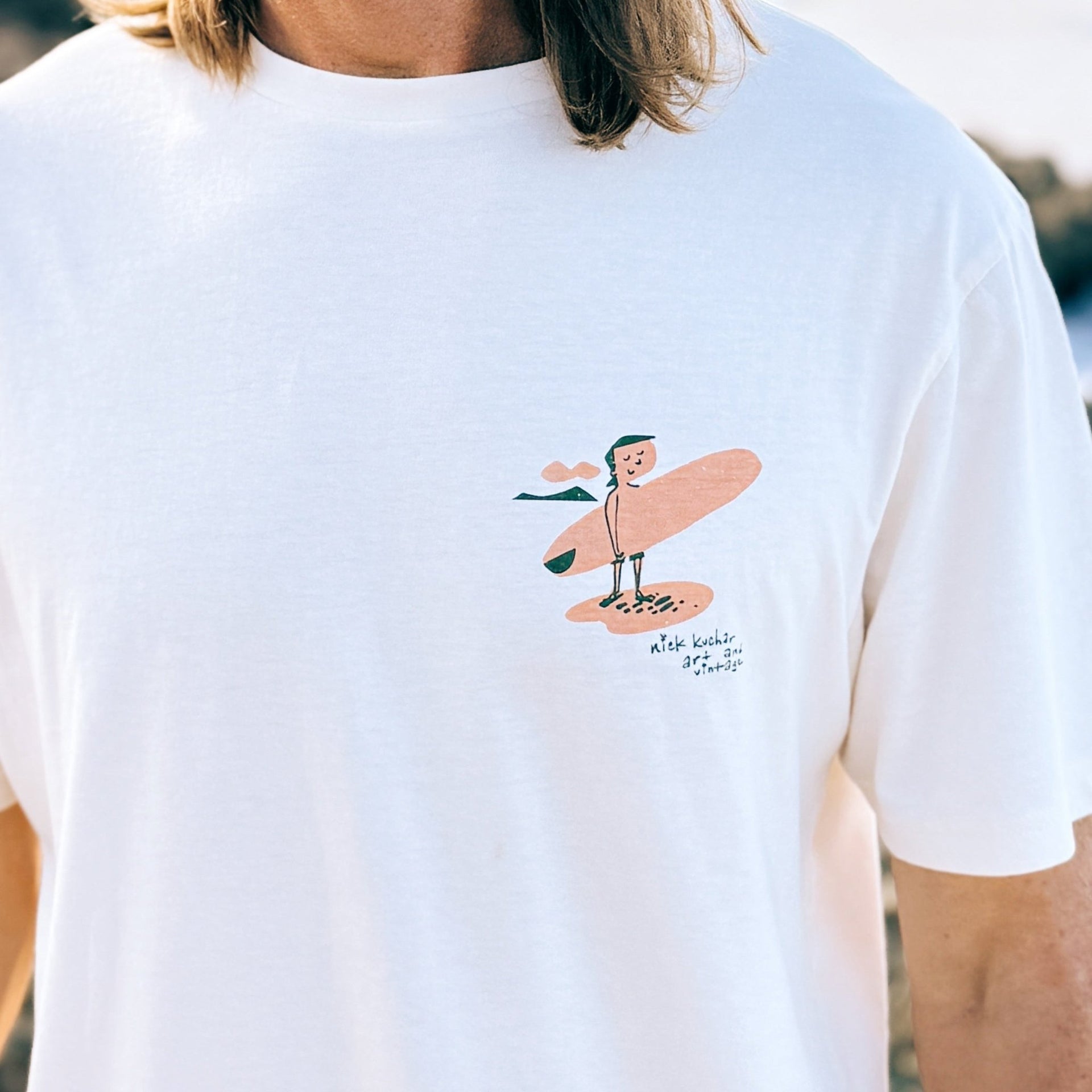 White t-shirt with a cartoon surf character holding a surfboard on the front, worn by a man.